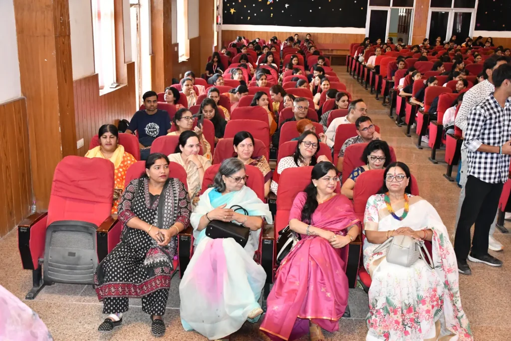 Audience seated in a large hall with red theater seats, many people facing forward during an event. Foreground shows women in colorful traditional clothing/patterned saris.