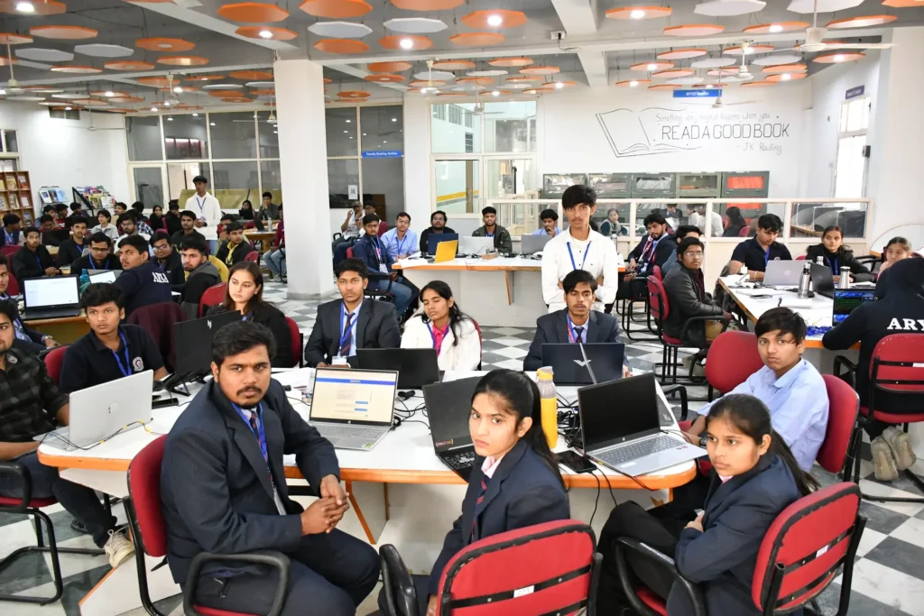 Large group of students in a computer lab working on laptops at round tables with orange ceiling decor overhead and a quote about reading on the wall in the background.