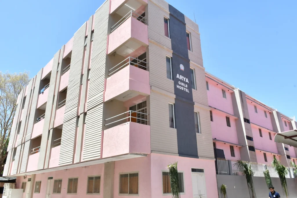 Front view of Arya Girls Hostel, a pink and gray multi-story building with balconies against a clear blue sky.